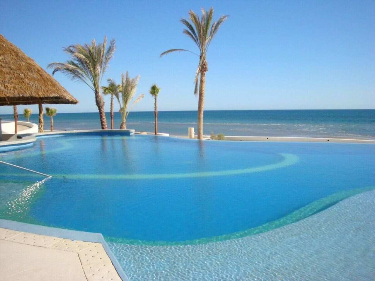 Una piscina d'aigua cristal·lina de color blau infinit, amb una estructura amb teulada de palla i palmeres.