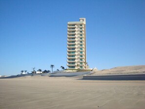 A tall, multi-story hotel with a flat roof, located on a sandy beach with palm trees in the background. - Amazing Beachfront Condo, Tessoro at Las Conchas, Puerto Penasco (Las Conchas)