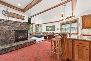 A spacious living area with a stone fireplace, a large sofa set, and a kitchen with wooden cabinets. - Monthly Lease - Granite Peak at Palisades - Tahoe Getaways - 4BR Dog Ok w Hot Tub & Shuttle (Olympic Valley)
