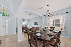 Un comedor con una mesa de madera dispuesta para cuatro personas, rodeada de sillas. Encima de la mesa hay una araña, y la habitación tiene un techo alto con un arco que da a otra estancia.