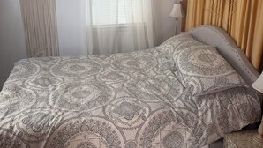 A bedroom with a bed, a bedside table, a hanging macrame wall decoration, a window with sheer curtains, and a wall-mounted lamp.