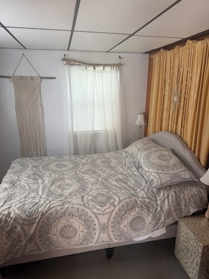 A bedroom with a bed, a bedside table, a hanging macrame wall decoration, a window with sheer curtains, and a wall-mounted lamp.