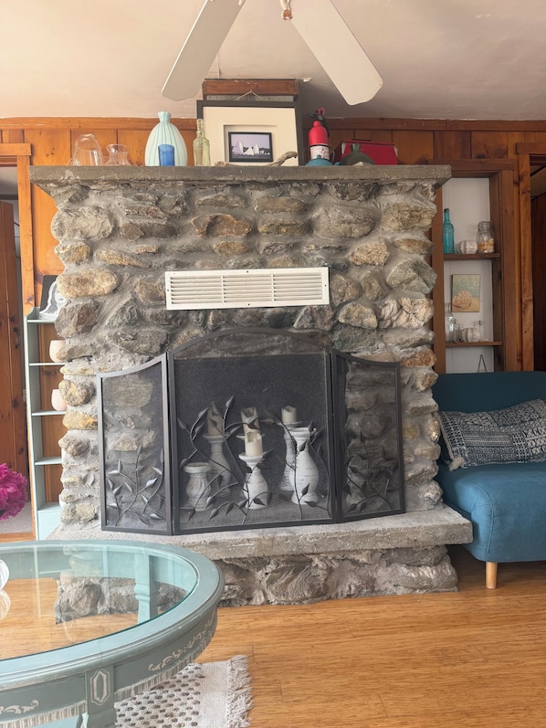 A stone fireplace with a framed picture above it, a glass table with a woven placemat, and a blue sofa in the foreground.