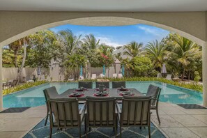 Un espace repas aménagé au bord de la piscine, avec une table dressée pour quatre personnes, entourée de chaises, et une vue sur un jardin tropical.