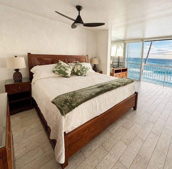 A large bed with a wooden frame, two bedside tables with lamps, a ceiling fan, and a view of the ocean through large windows.