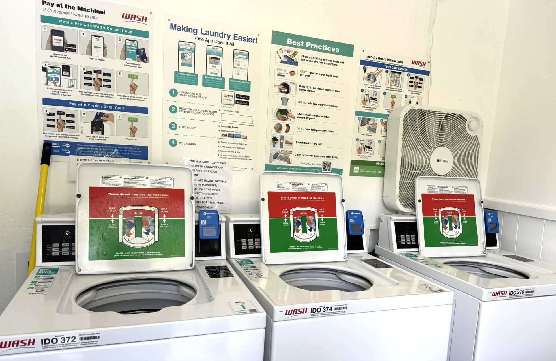 A laundromat with multiple washing machines, a fan, and instructional posters on payment methods and laundry practices.