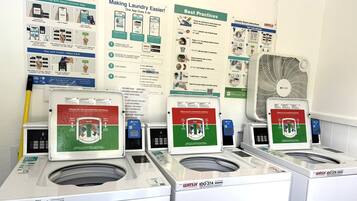 A laundromat with multiple washing machines, a fan, and instructional posters on payment methods and laundry practices.