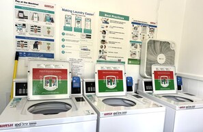 A laundromat with multiple washing machines, a fan, and instructional posters on payment methods and laundry practices.