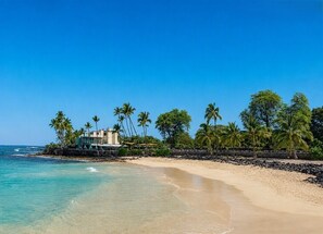 A beach with clear blue water, a sandy shore, and a building with a pool surrounded by palm trees.