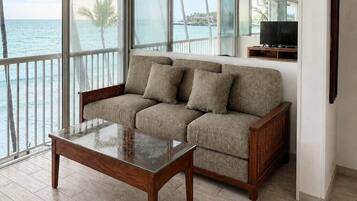 A modern living room with a sofa and a glass coffee table, large windows with a view of the sea, and a flat-screen TV on a wooden stand.