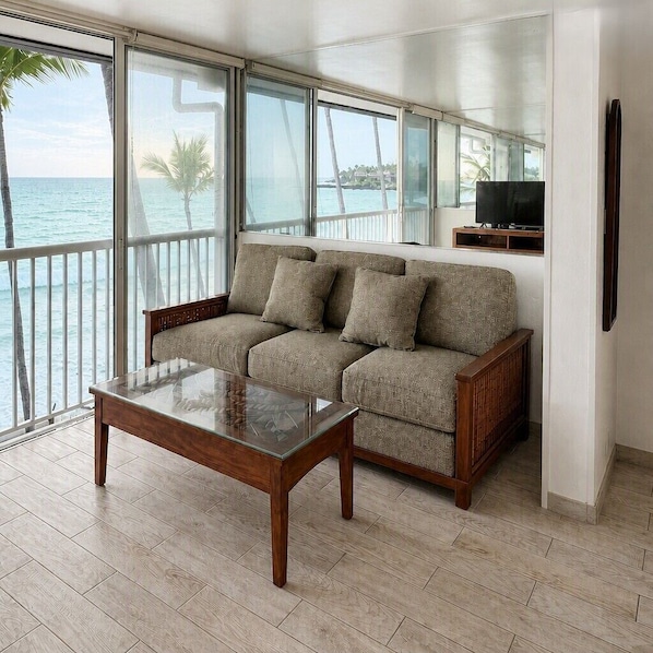 A modern living room with a sofa and a glass coffee table, large windows with a view of the sea, and a flat-screen TV on a wooden stand.