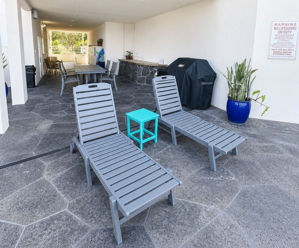 A covered outdoor seating area with lounge chairs and a small table, a barbecue grill, and a potted plant.