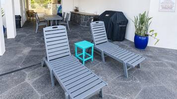 A covered outdoor seating area with lounge chairs and a small table, a barbecue grill, and a potted plant.