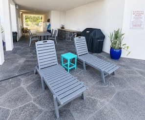A covered outdoor seating area with lounge chairs and a small table, a barbecue grill, and a potted plant. - KONA MAGIC SANDS 314 - Top Floor Oceanfront Corner Penthouse - Magic Sands Beach (Kailua-Kona)