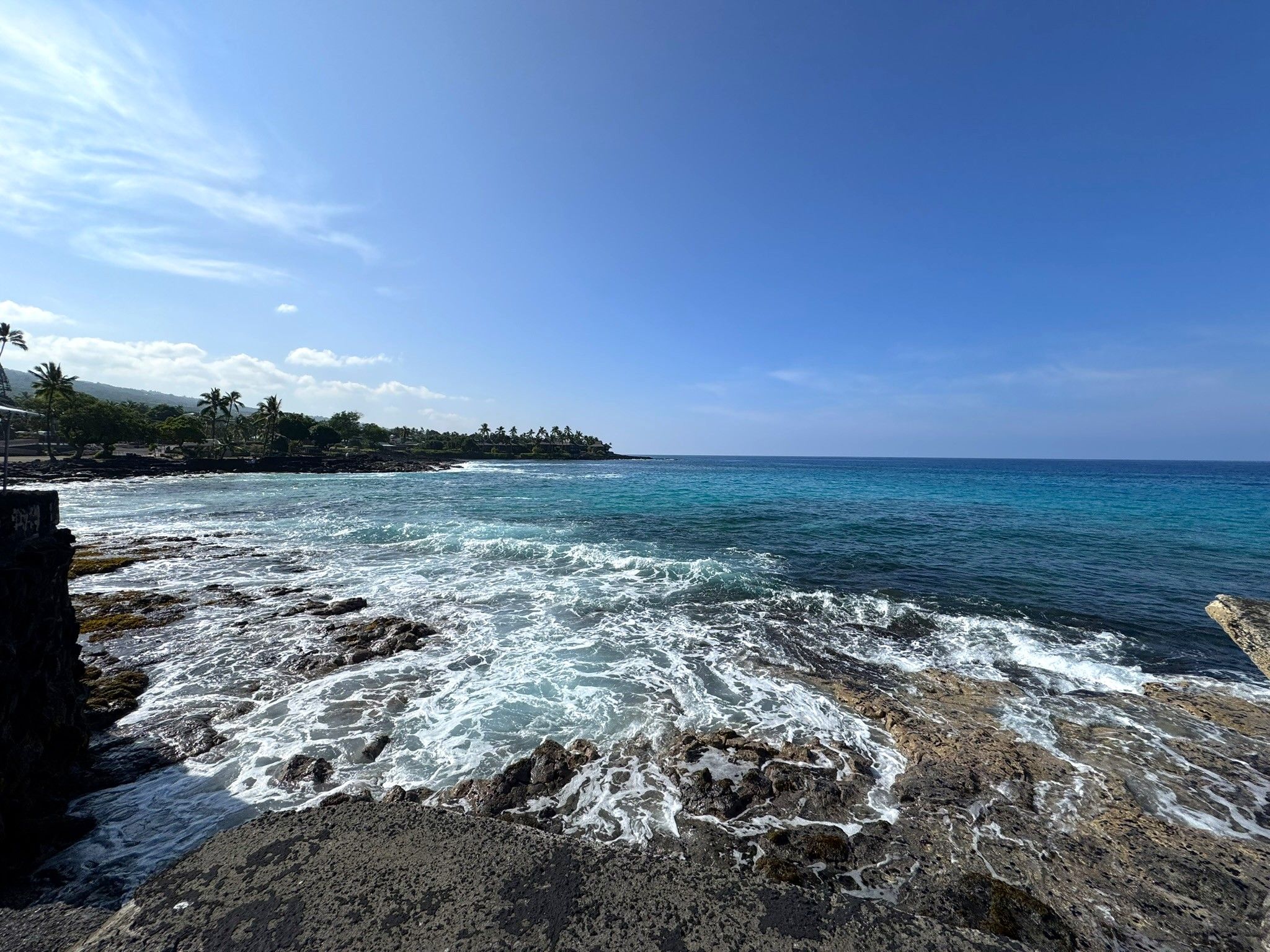 A rocky shoreline with waves crashing onto the rocks, a clear blue sky, and a distant island with palm trees.