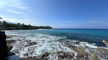 A rocky shoreline with waves crashing onto the rocks, a clear blue sky, and a distant island with palm trees.