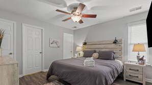 Ein Schlafzimmer mit Bett, Deckenventilator, Nachttisch, Lampe, Teppich und einer Tür.
