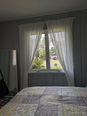 A bedroom with a bed, a window with curtains, a radiator, and a mirror.