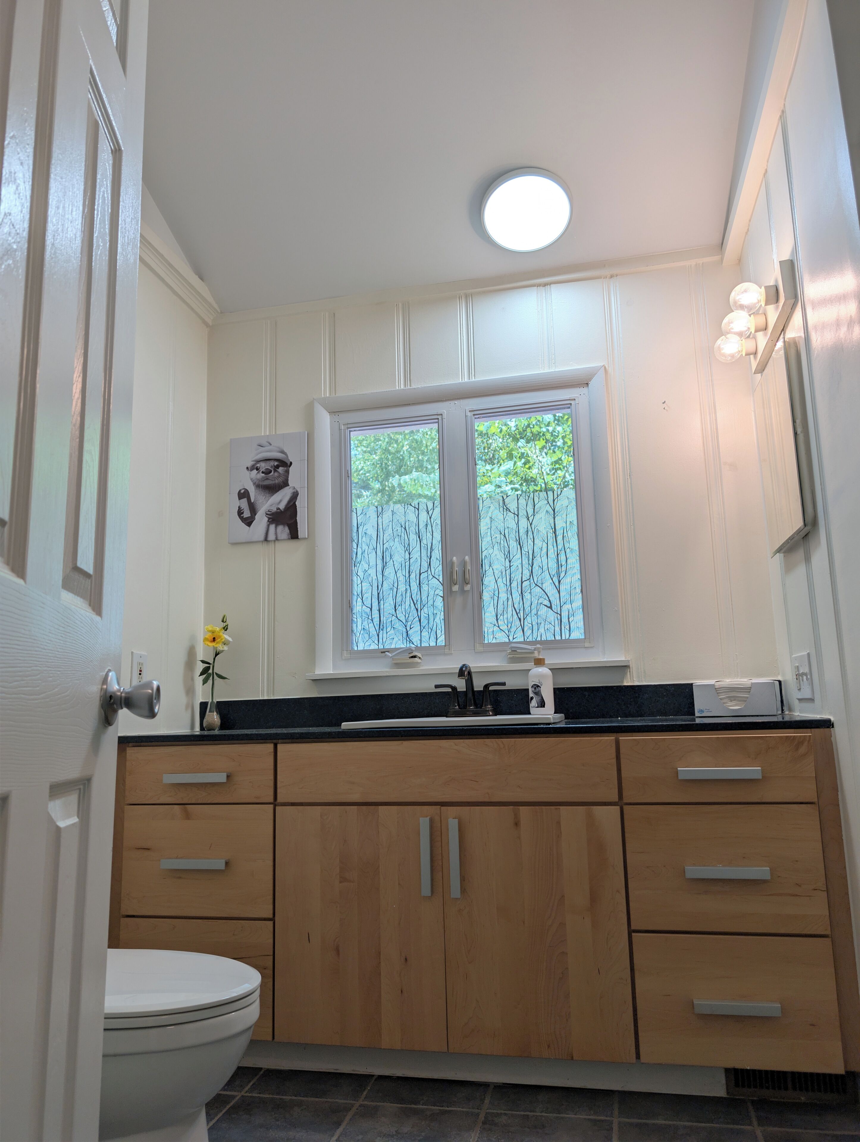 Une salle de bain moderne avec des toilettes blanches, des meubles en bois, un comptoir noir, une fenêtre donnant sur les arbres et un luminaire fixé au mur.