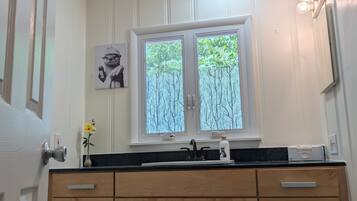 Une salle de bain moderne avec des toilettes blanches, des meubles en bois, un comptoir noir, une fenêtre donnant sur les arbres et un luminaire fixé au mur.