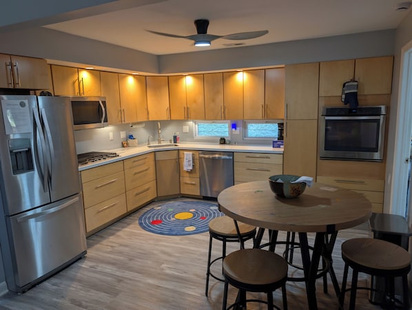 A modern kitchen with wooden cabinets, stainless steel appliances, a round dining table with stools, and a ceiling fan.