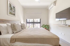 Une chambre à coucher avec un lit, des oreillers, une couverture, une plante en pot, un téléviseur et une fenêtre donnant sur des immeubles.
