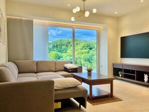 Ein modernes Wohnzimmer mit Sofa, Couchtisch, Fernseher und einem großen Fenster mit Blick auf einen Wald.