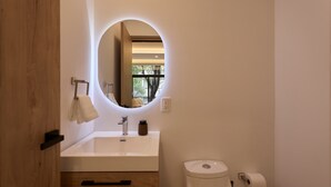 Un baño moderno con tocador de madera, un espejo redondo con iluminación, inodoro y cubo de basura.
