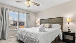 Un dormitorio con una cama grande, un ventilador de techo, una mesita de noche con lámpara y una ventana con cortinas.