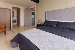Ein Hotelzimmer mit einem großen Bett, einem Sessel, einem Nachttisch sowie Blick ins Bad und zur Küche.