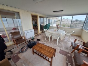A room with a large window, a table with chairs, a wooden bench, a radiator, and a vacuum cleaner.