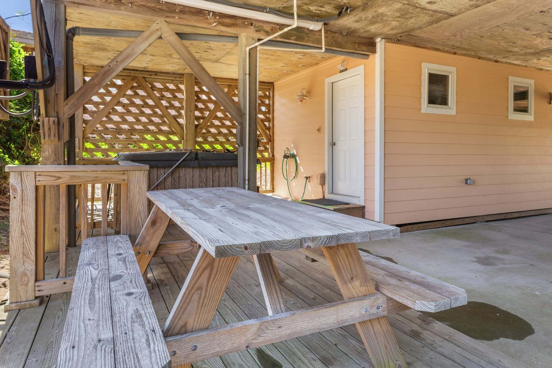 A wooden picnic table and bench under a wooden pergola with a door and windows in the background.
