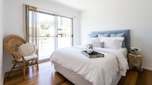 Une chambre à coucher avec un grand lit, une chaise en osier, une table de chevet et une vue sur l’extérieur, accessible par une porte-fenêtre coulissante.