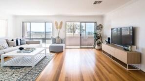 Ein modernes Wohnzimmer mit einer Couch, einem Couchtisch und einem Fernseher, großen Fenstern und einem Balkon.