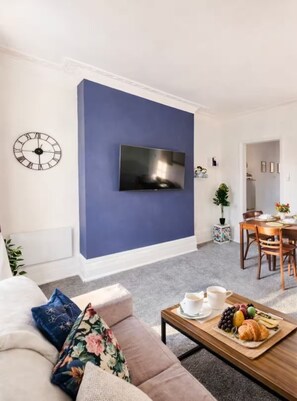 Ein Wohnzimmer mit blauer Akzentwand, einem Flachbildfernseher, einer Couch, einem Couchtisch mit Obst und einem Croissant sowie einem Essbereich mit Stühlen und Tisch.