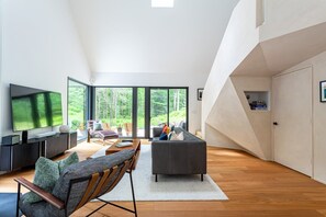 Een moderne woonkamer met een grote televisie, comfortabele zitmogelijkheden en uitzicht op een bos door grote ramen.