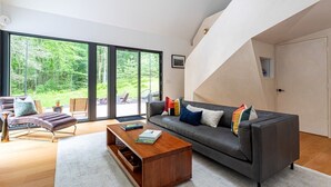 Una sala de estar moderna con un sofá, un sillón y una mesa de centro, ventanas amplias y una escalera.