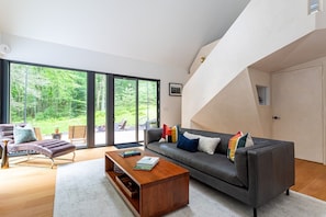 Een moderne woonkamer met een bank, fauteuil, salontafel, grote ramen en een trap.