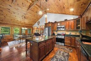 A spacious kitchen with wooden cabinets, a large island, stainless steel appliances, and a dining area with a table and chairs. - Spacious Cabin Retreat with Hot Tub & Game Room (Sugar Grove)