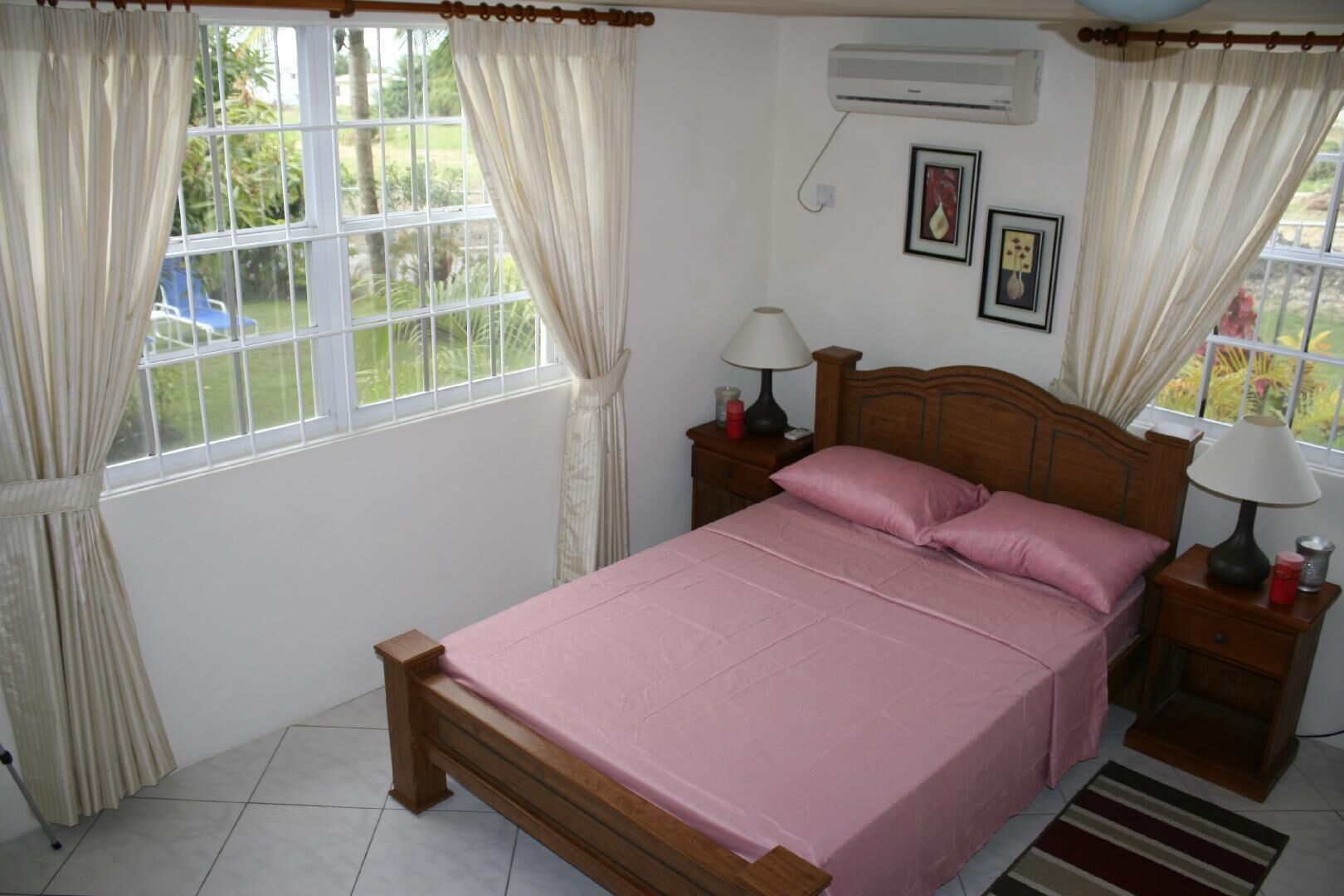 A bedroom with a bed, two bedside tables with lamps, a window with curtains, and an air conditioning unit.