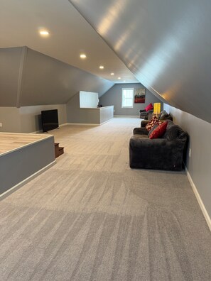 A spacious attic room with a high ceiling, a built-in entertainment unit, a large window, and a cozy seating area.