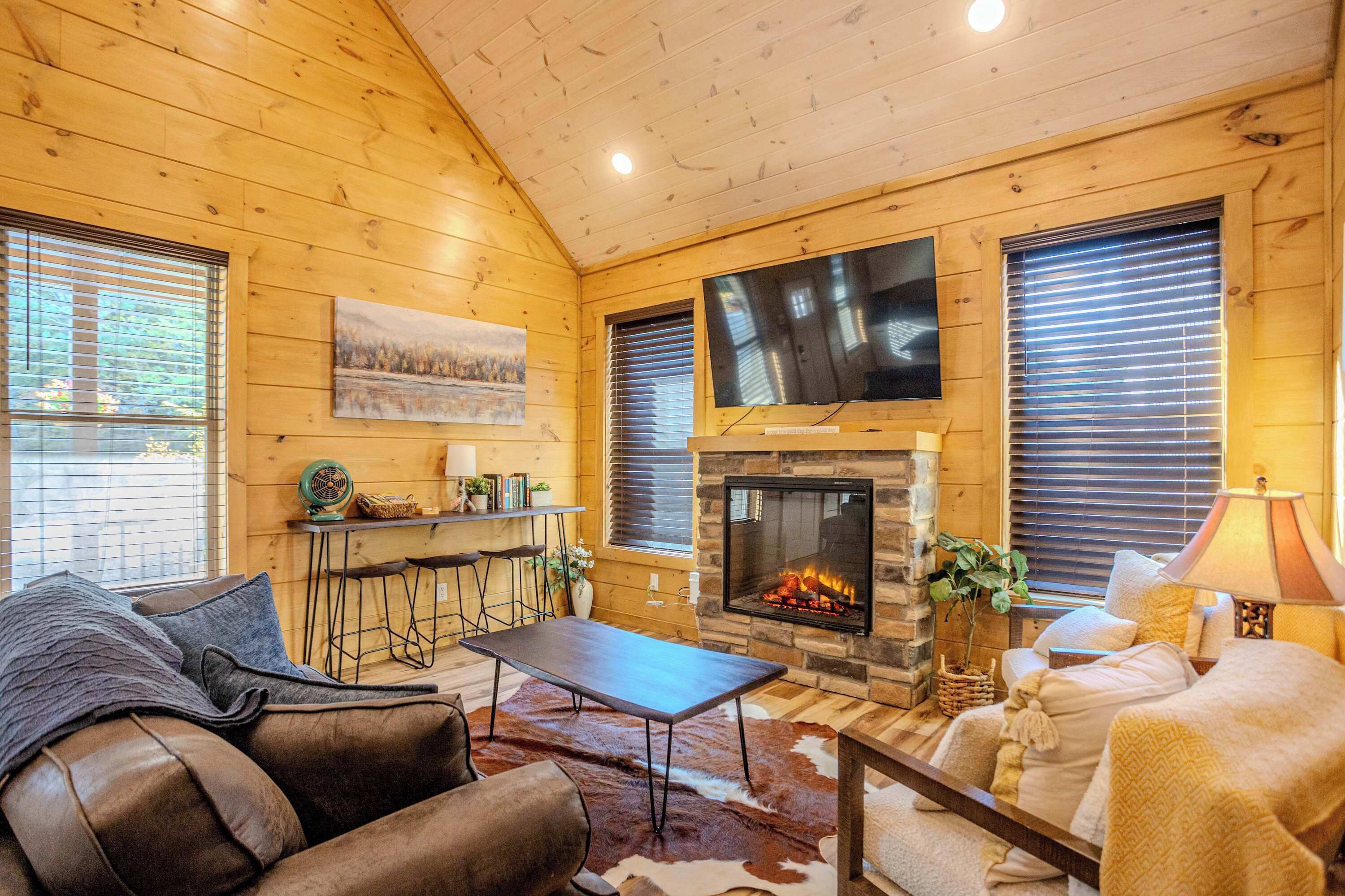 A cozy cabin interior with a stone fireplace, wooden walls, a leather sofa, and a dining area.