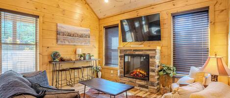 A cozy cabin interior with a stone fireplace, wooden walls, a leather sofa, and a dining area.