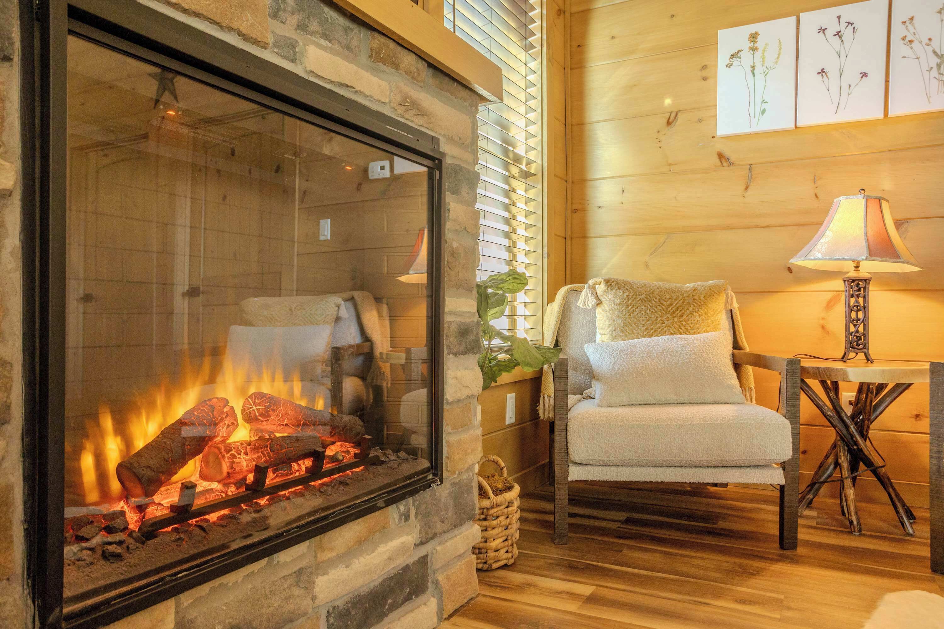 A fireplace with logs, a chair, a lamp, and framed botanical artwork on the wall.