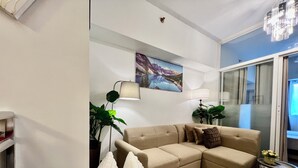 Ein Wohnzimmer mit einem beigefarbenen Ecksofa, zwei Stehlampen, einem großen Gemälde und einer Glastür, die auf einen Balkon führt.
