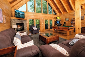 A log cabin interior with a fireplace, a pool table, and a large window overlooking greenery.