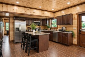 A kitchen with wooden paneling, stainless steel appliances, a central island with bar stools, and a view of greenery outside. - Cloud's Rest | Luxury Estate • Pool & Pavilion (Sevierville)