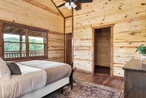 A wooden cabin bedroom with a bed, a ceiling fan, a window with a view, and a door. - Cloud's Rest | Luxury Estate • Pool & Pavilion (Sevierville)