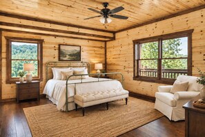 A wooden cabin bedroom with a bed, bedside tables, a chair, and a view of the outdoors. - Cloud's Rest | Luxury Estate • Pool & Pavilion (Sevierville)