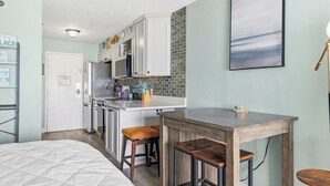 Een kleine keuken met een centraal eiland, een eethoek met krukken, en een bed met een gewatteerd dekbed.